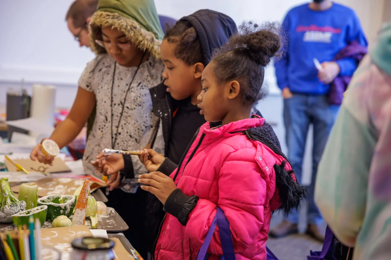 Three children focus intently on a hands-on science activity, using paint and vegetables as stamps to create art. The table in front of them is covered with colorful supplies and paint-covered vegetables, while adults look on in the background.