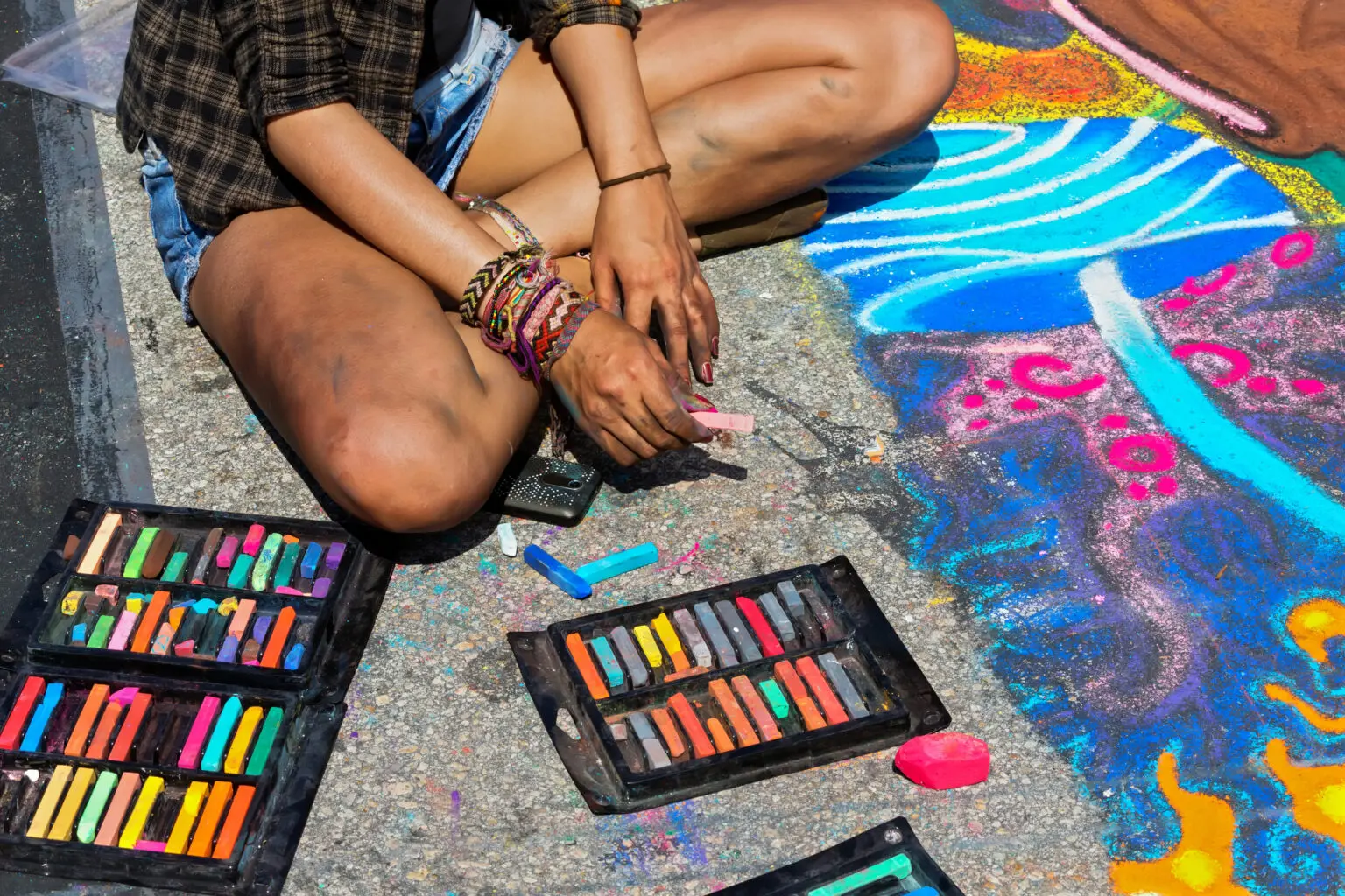 Young woman sitting on the sidewalk with her legs crossed. Surrounded by pastel chalk art and sidewalk chalk.